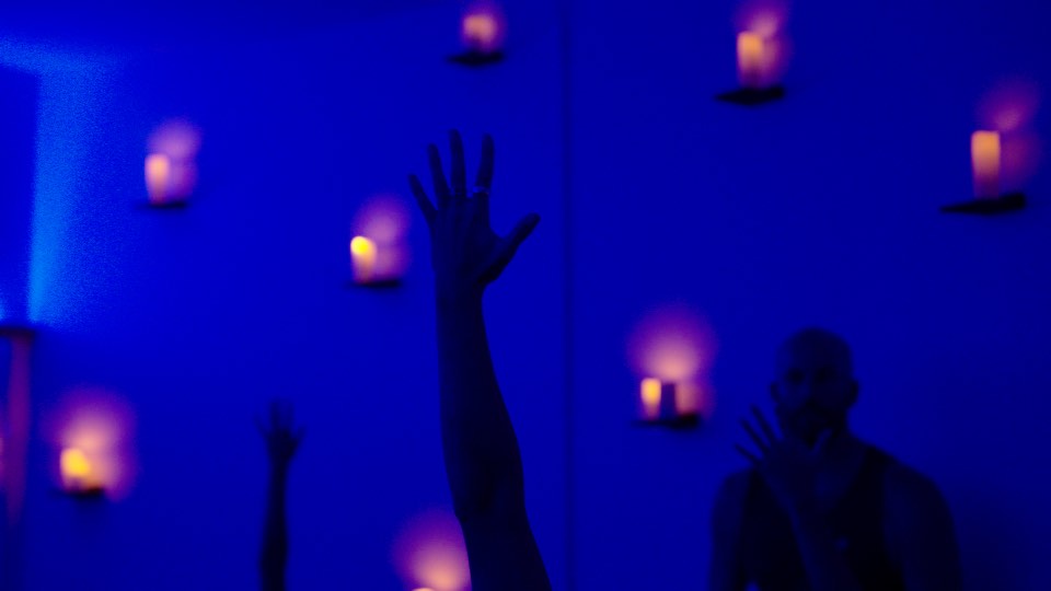 A raised hand in silhouette against a blue-lit wall lined with candles, in the Waialua yoga room