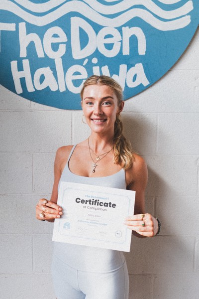 A graduate holding her certificate of completion under the Den Haleʻiwa sign