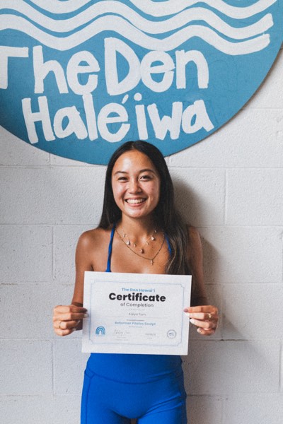 A graduate holding her certificate of completion under the Den Haleʻiwa sign
